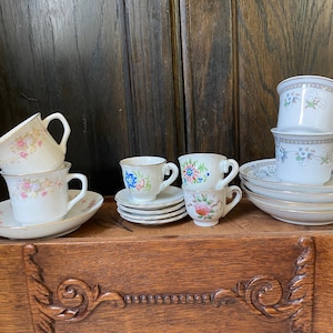 Puede incluir: Una colección de tazas de té y platillos vintage con diseños florales. Las tazas y los platillos son blancos con ribetes dorados y presentan delicados diseños florales rosas, azules y amarillos. El conjunto incluye varios tamaños y estilos, dispuestos sobre una superficie de madera.