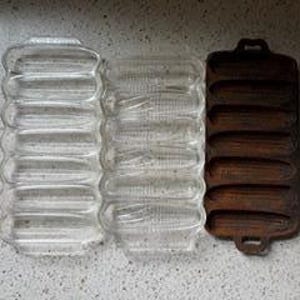 Puede incluir: Tres moldes de horneado rectangulares dispuestos uno al lado del otro. Dos moldes son de vidrio transparente con hendiduras en forma de maíz, y un molde es de hierro fundido marrón oscuro con cavidades idénticas y dos asas.