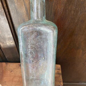 Puede incluir: Una botella de vidrio transparente con una superficie texturizada. La botella tiene un cuello redondeado y un cuerpo cuadrado. La botella est&aacute; vac&iacute;a y tiene una imagen tenue de una cabeza humana en la parte delantera.