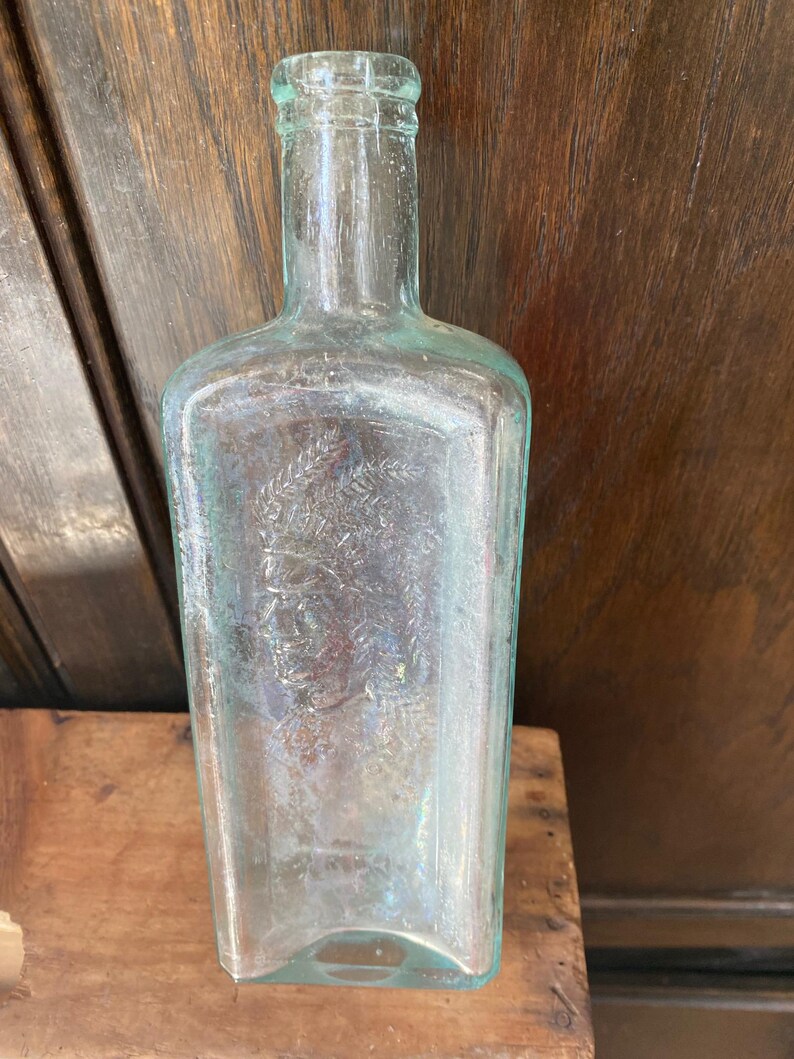 Puede incluir: Una botella de vidrio transparente con una superficie texturizada. La botella tiene un cuello redondeado y un cuerpo cuadrado. La botella est&aacute; vac&iacute;a y tiene una imagen tenue de una cabeza humana en la parte delantera.