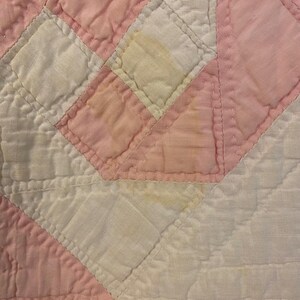 May include: A quilted fabric textile featuring a geometric pattern of alternating white and light pink squares and triangles. The surface displays visible stitching lines throughout the fabric, with some areas showing light yellow discolouration.