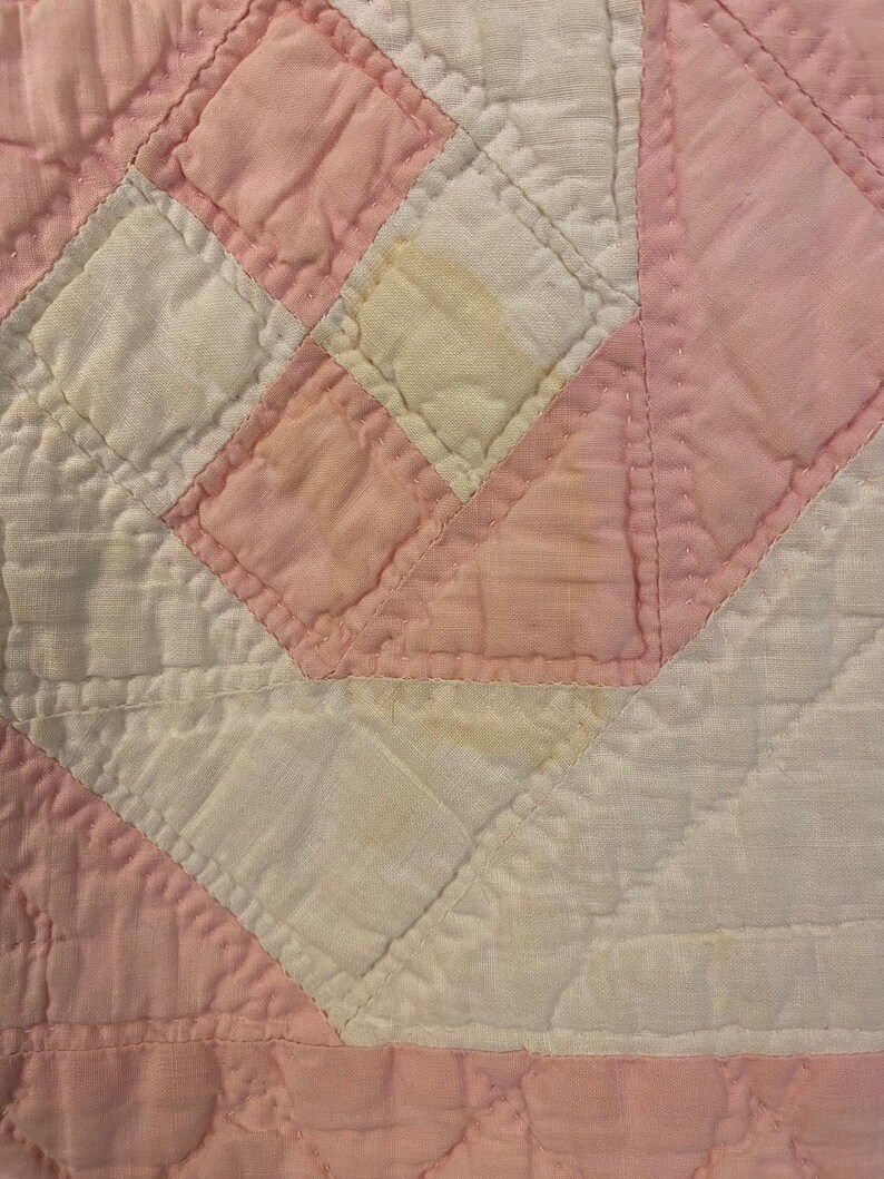 May include: A quilted fabric textile featuring a geometric pattern of alternating white and light pink squares and triangles. The surface displays visible stitching lines throughout the fabric, with some areas showing light yellow discolouration.
