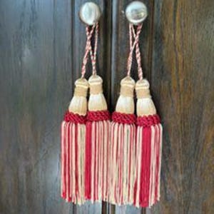 May include: Two decorative tassels hanging from silver-toned knobs. Each tassel features a braided cord in red and white, a beige cap, a thick red braided band, and a long fringe composed of alternating strands of cream and deep red thread.