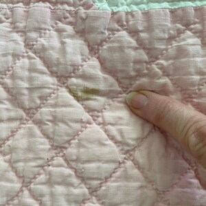 May include: A light pink quilted fabric featuring a diamond-stitched pattern and a white section at the top edge. A small, brownish discoloration is visible on the pink surface, highlighting a specific area of the textile material.