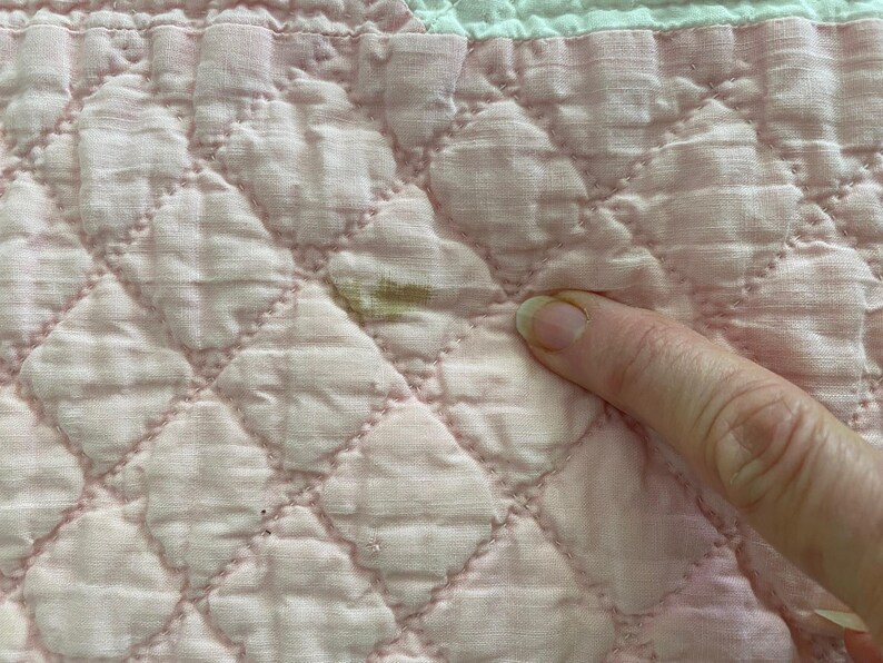 May include: A light pink quilted fabric featuring a diamond-stitched pattern and a white section at the top edge. A small, brownish discoloration is visible on the pink surface, highlighting a specific area of the textile material.