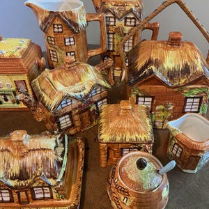 Op de afbeelding: Een verzameling keramische theepotten en serveerstukken in de vorm van huisjes. De set bevat een theepot, een melkkan, een suikerpot, een serveerschaal en andere bakjes. De stukken zijn bruin met rieten daken en raamdetails.
