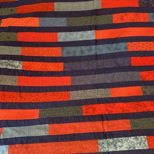 May include: A handmade quilt with a patchwork design. The quilt features horizontal stripes of navy blue, red, gray, and patterned fabrics. The red fabrics have various patterns. The quilt is a cozy and decorative textile.