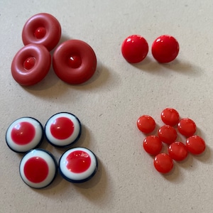 May include: A collection of red buttons in various shapes and sizes. There are three donut-shaped buttons, two round buttons, four buttons with a red center and white and blue border, and a cluster of small, round buttons.