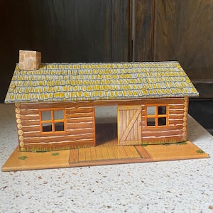 Può includere: Una casetta di legno giocattolo vintage con tetto giallo, cornici delle finestre arancioni e una porta di legno. La casetta è composta da pezzi marroni a forma di tronco e poggia su una base beige. Un piccolo camino è visibile sul tetto.