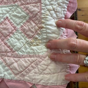 May include: A quilted fabric piece featuring a geometric pattern in white and pale pink. The textile has a scalloped pink border and shows visible brown staining on the white fabric sections, indicating age and wear on the material.