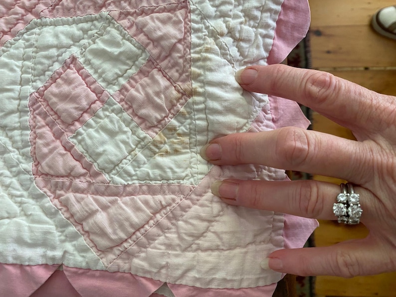 May include: A quilted fabric piece featuring a geometric pattern in white and pale pink. The textile has a scalloped pink border and shows visible brown staining on the white fabric sections, indicating age and wear on the material.