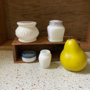 May include: A selection of vintage white milk glass jars and containers, including a decorative jar with floral relief, a jar with a metal lid, and a small jar. A yellow pear and a small tin of Silicote Line Dressing are also present.