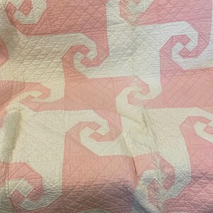 May include: A quilt featuring a repeating geometric pattern of interlocking white and light pink spiral shapes. The fabric has a diamond-stitched quilted texture throughout, and the top edge is finished with a scalloped pink border.