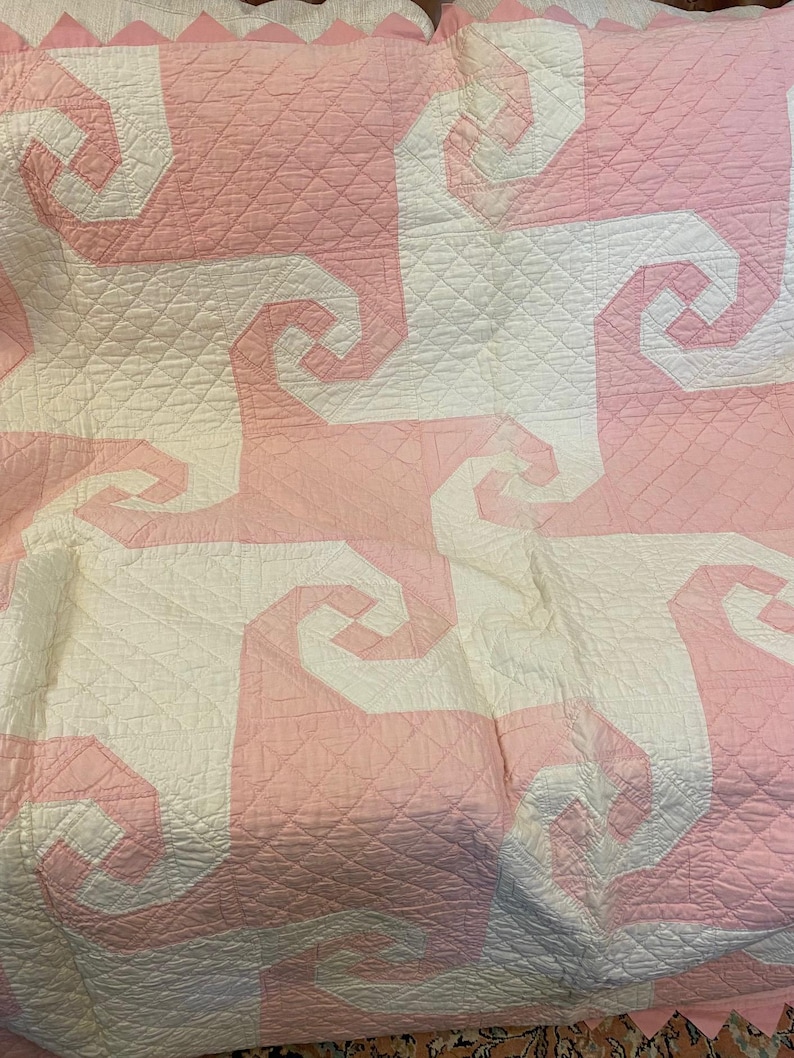May include: A quilt featuring a repeating geometric pattern of interlocking white and light pink spiral shapes. The fabric has a diamond-stitched quilted texture throughout, and the top edge is finished with a scalloped pink border.