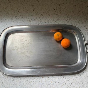 May include: A rectangular silver-toned metal serving tray with rounded corners and two side handles featuring dark brown accents. Two bright orange citrus fruits are positioned on the flat surface of the tray, which has a smooth metallic finish.