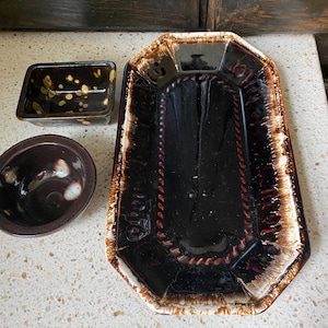 May include: A collection of ceramic serving dishes in shades of brown and black. The set includes a rectangular tray with a decorative border, a small rectangular dish with gold accents, and a round bowl.