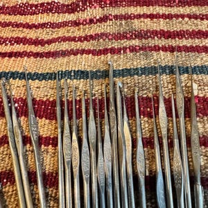 May include: A collection of vintage crochet hooks made of silver metal. The hooks are arranged in a row, with varying sizes and markings. The background is a colorful, patterned textile.