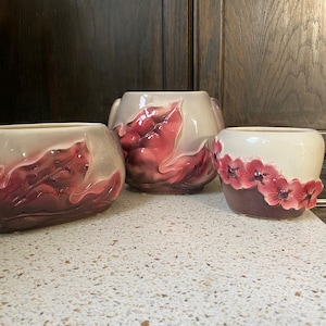May include: Three ceramic flower pots with pink and white floral designs. The pots are different sizes and shapes.