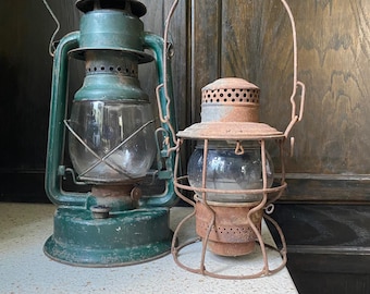 May include: Two vintage lanterns. One is a larger, green metal lantern with a clear glass globe and a wire handle. The other is a smaller, rusty metal lantern with a clear glass globe and a wire handle. Both are on a white surface.