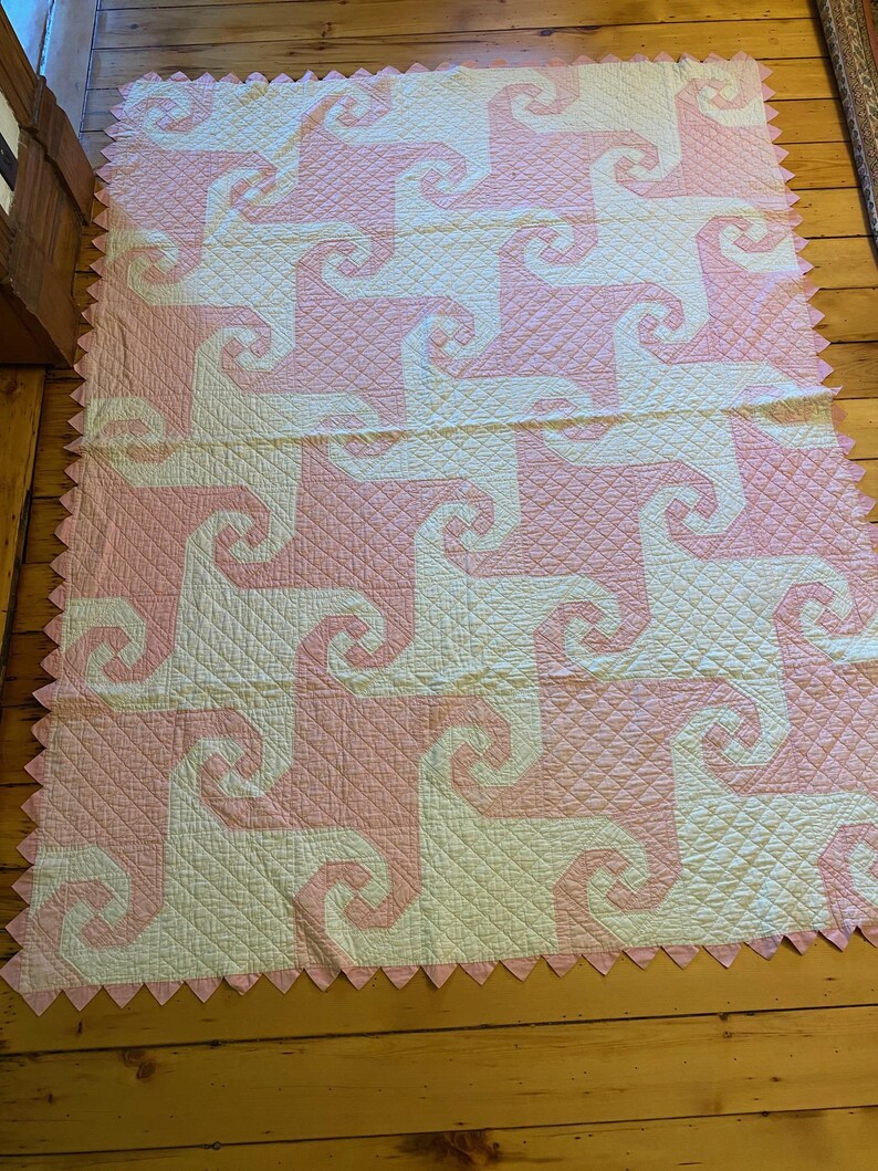 May include: A rectangular quilt featuring a repeating spiral pattern in alternating light pink and cream fabric. The surface has a diamond-shaped quilted texture throughout, and the edges are finished with a pink sawtooth border.