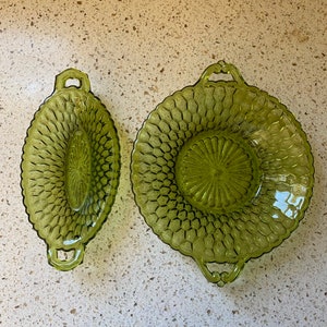 May include: Two green glass serving dishes with handles. The larger dish is round and the smaller dish is oval. Both dishes have a textured, raised pattern.