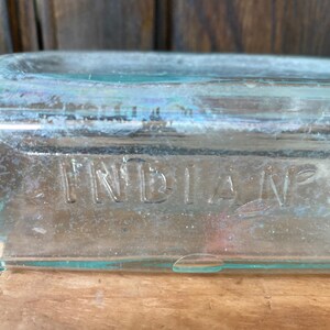 Puede incluir: Una botella de vidrio transparente con la palabra "INDIAN" grabada en el lateral.