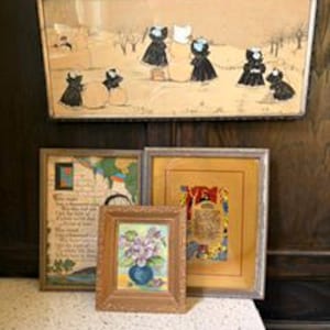 May include: A collection of framed art pieces, including a large rectangular piece depicting figures in black dresses, and smaller square and rectangular pieces with floral and abstract designs. The frames vary in style and color.