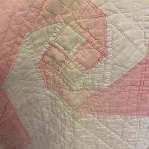 May include: A quilted textile featuring a geometric pinwheel pattern in alternating shades of soft pink and white. The fabric displays a textured, stitched surface with a repeating square design, showcasing intricate needlework and fabric piecing.