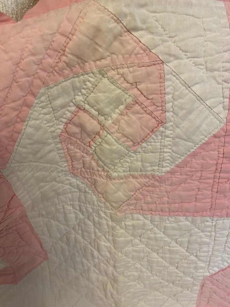 May include: A quilted textile featuring a geometric pinwheel pattern in alternating shades of soft pink and white. The fabric displays a textured, stitched surface with a repeating square design, showcasing intricate needlework and fabric piecing.