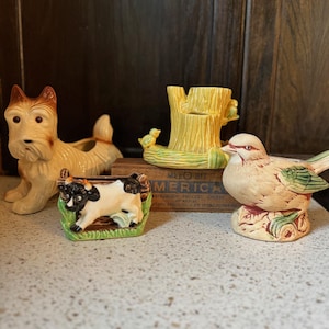 May include: A selection of vintage ceramic ornaments. The collection includes a tan and brown terrier, a yellow tree stump with a bird, a cow, and a bird perched on a base. The figurines are displayed on a speckled countertop.
