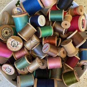 Puede incluir: Un bol lleno de carretes de hilo de varios colores, incluyendo rojo, azul, verde, marrón, rosa y beige. Los carretes son de madera y tienen etiquetas con el texto "J&P. Coats" y "Coats".