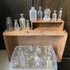 Puede incluir: Colección de botellas y tarros de vidrio antiguos de varias formas y tamaños. Algunas botellas se exhiben en una estantería de madera, mientras que otras están en un recipiente de plástico transparente. Las botellas son transparentes y muestran signos de edad.