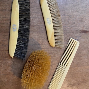 May include: A collection of vintage grooming tools, including two rectangular brushes with dark bristles, a round brush, and a comb. The brushes have light-colored handles with a blue emblem. The items are arranged on a wooden surface.
