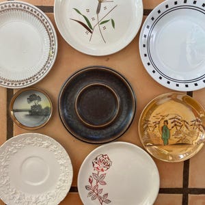 Puede incluir: Una colección de varios platos decorativos. Los platos presentan diversos diseños, incluyendo patrones florales, bordes geométricos y escenas de paisajes. Los colores incluyen blanco, marrón, negro y dorado. Los platos están dispuestos sobre una superficie de baldosas marrones.