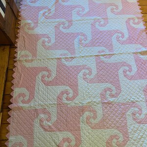 May include: A rectangular quilt featuring a repeating geometric pattern of interlocking pink and white spiral shapes. The fabric surface displays a fine diamond-shaped quilted texture throughout, and the edges are finished with a pink sawtooth border.