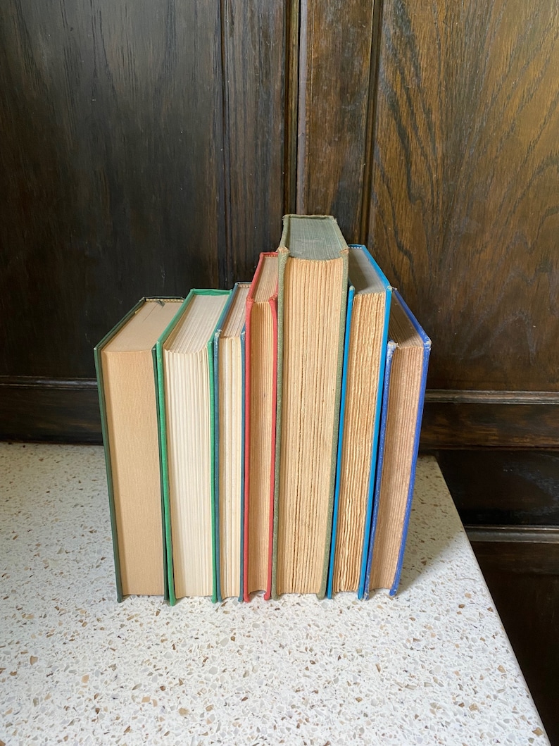 K&ouml;nnte beinhalten: Ein Stapel von sieben alten gebundenen B&uuml;chern mit verschiedenen R&uuml;ckenfarben. Die R&uuml;cken sind gr&uuml;n, creme, blau, rot, beige, blau und blau. Die B&uuml;cher sind in einer Reihe auf einer wei&szlig;en Oberfl&auml;che angeordnet.