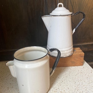 May include: Two white enamelware coffee pots with black handles and trim. One pot has a lid. The pots are on a wooden block and a speckled countertop. The background is a dark wooden cabinet.