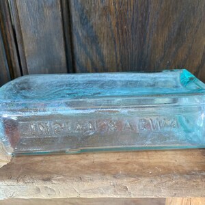 Puede incluir: Una botella vintage de vidrio transparente con un tono azul. La botella tiene una forma rectangular y las palabras "INDIANAS AGWA" est&aacute;n grabadas en el vidrio.