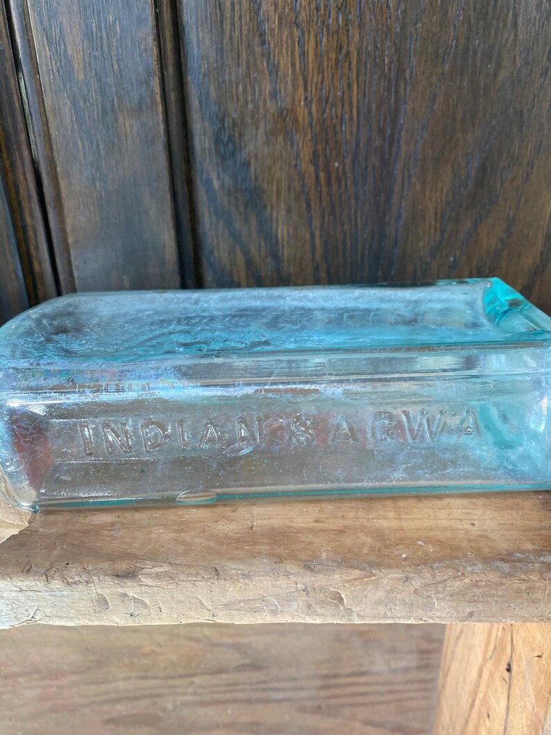 Puede incluir: Una botella vintage de vidrio transparente con un tono azul. La botella tiene una forma rectangular y las palabras "INDIANAS AGWA" est&aacute;n grabadas en el vidrio.