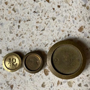 Puede incluir: Tres pesas antiguas de latón de diferentes tamaños. La pesa más pequeña está marcada con "10 G.M.", la del medio con "20" y la más grande con "2 oz". Las pesas son circulares y tienen un borde ligeramente elevado.
