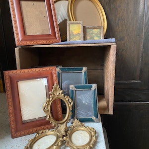 以下が含まれることがあります： さまざまな形とサイズのヴィンテージ写真フレームのコレクション。フレームには、茶色、金、ティール色の長方形、楕円形、装飾的なデザインが含まれます。一部のフレームは空で、他のフレームには小さな風景画が含まれています。
