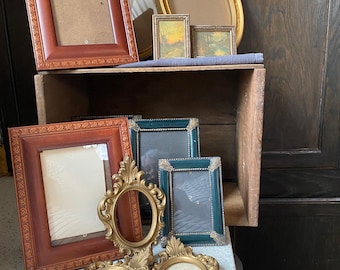 May include: A collection of vintage picture frames in various shapes and sizes. Frames include rectangular, oval, and ornate designs in shades of brown, gold, and teal. Some frames are empty, while others contain small landscape paintings.