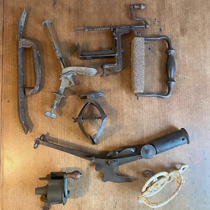 May include: A collection of vintage tools, including a metal hand crank, a metal clamp, a metal scraper, a metal handle with a wooden roller, a metal handle with a metal blade, a metal handle with a metal hook, a metal handle with a metal hook, and a white metal handle with a metal hook.