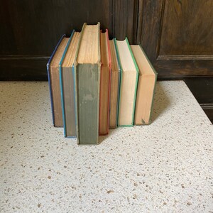 K&ouml;nnte beinhalten: Ein Stapel von sechs alten gebundenen B&uuml;chern mit unterschiedlichfarbigen R&uuml;ckseiten. Die B&uuml;cher sind vertikal angeordnet, wobei die R&uuml;ckseiten zum Betrachter zeigen. Die B&uuml;cher befinden sich auf einer wei&szlig;en Oberfl&auml;che mit einem gesprenkelten Muster.