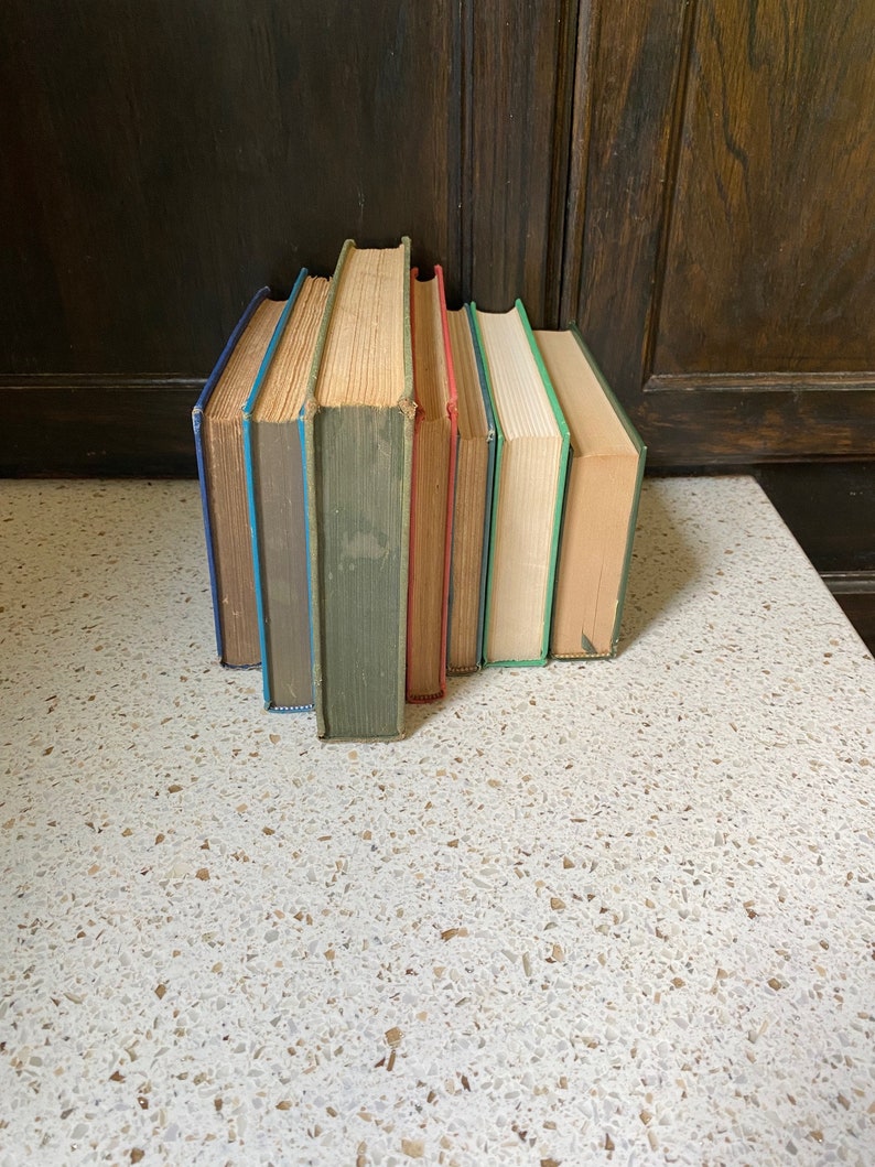 K&ouml;nnte beinhalten: Ein Stapel von sechs alten gebundenen B&uuml;chern mit unterschiedlichfarbigen R&uuml;ckseiten. Die B&uuml;cher sind vertikal angeordnet, wobei die R&uuml;ckseiten zum Betrachter zeigen. Die B&uuml;cher befinden sich auf einer wei&szlig;en Oberfl&auml;che mit einem gesprenkelten Muster.