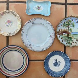 Puede incluir: Una colección de platos decorativos y una bandeja pequeña. Los platos presentan varios diseños, incluyendo patrones florales, bordes a rayas y un paisaje. Los colores incluyen azul, blanco, dorado, rojo y verde. La bandeja es azul claro con un diseño floral.