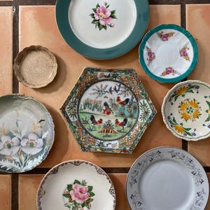 May include: A collection of decorative plates with floral and geometric designs. The plates vary in size, shape, and color, including a green-rimmed plate with a pink flower, a hexagonal plate with roosters, and plates with gold trim.