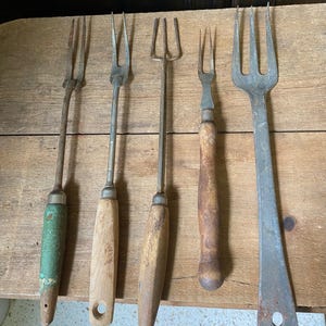 Op de afbeelding: Vijf vintage metalen vorken met houten handvatten. De handvatten variëren in kleur en vorm, waarvan één groen geverfd. De vorken hebben drie of vier tanden en vertonen tekenen van slijtage, wat duidt op antiek keukengereedschap.