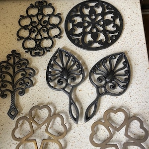 Puede incluir: Una colección de salvamanteles decorativos en varias formas y diseños. Los salvamanteles incluyen un salvamanteles redondo con estampado floral, un salvamanteles en forma de árbol y salvamanteles en forma de corazón. Los salvamanteles son de metal y de colores negro y bronce.