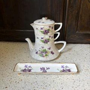 Puede incluir: Un juego de té de cerámica blanca para una persona, que incluye una tetera y una taza apilables con un patrón de violetas y borde dorado. El conjunto cuenta con una bandeja rectangular a juego decorada con las mismas flores violetas.